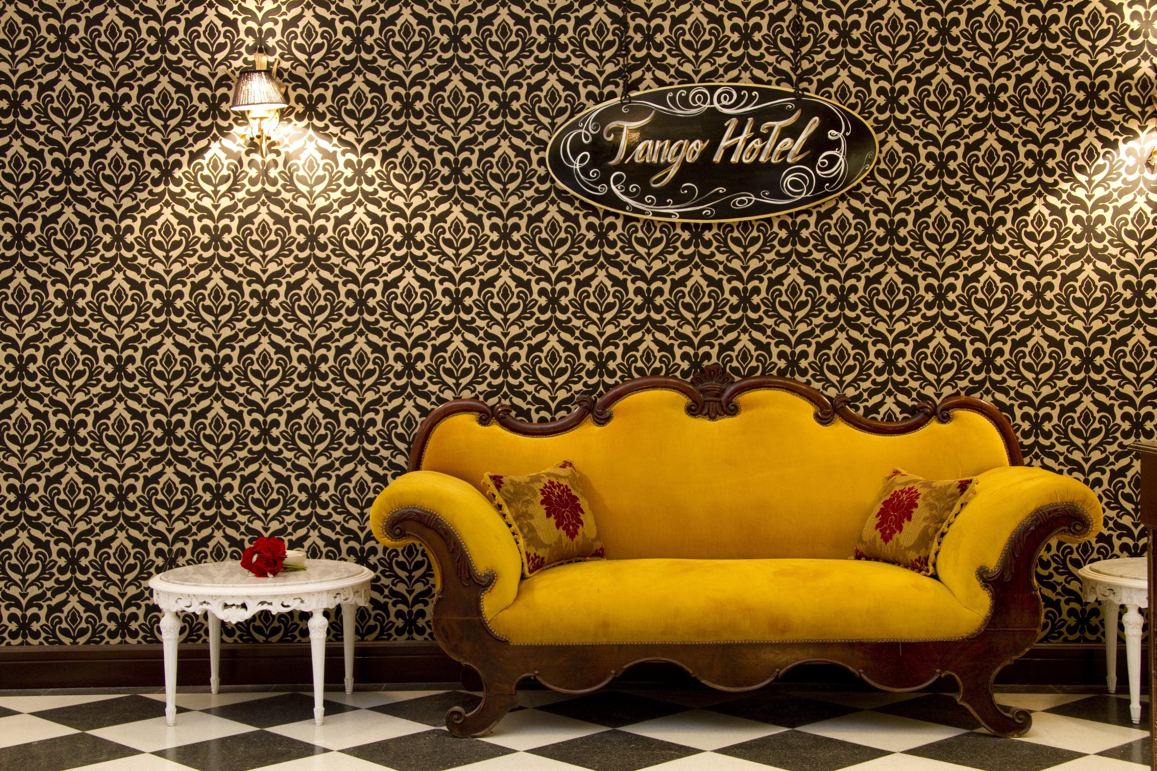 A yellow sofa with ornate wooden detailing, a white round table with a red rose, and a patterned wallpaper with a sign reading "Tango Hotel".