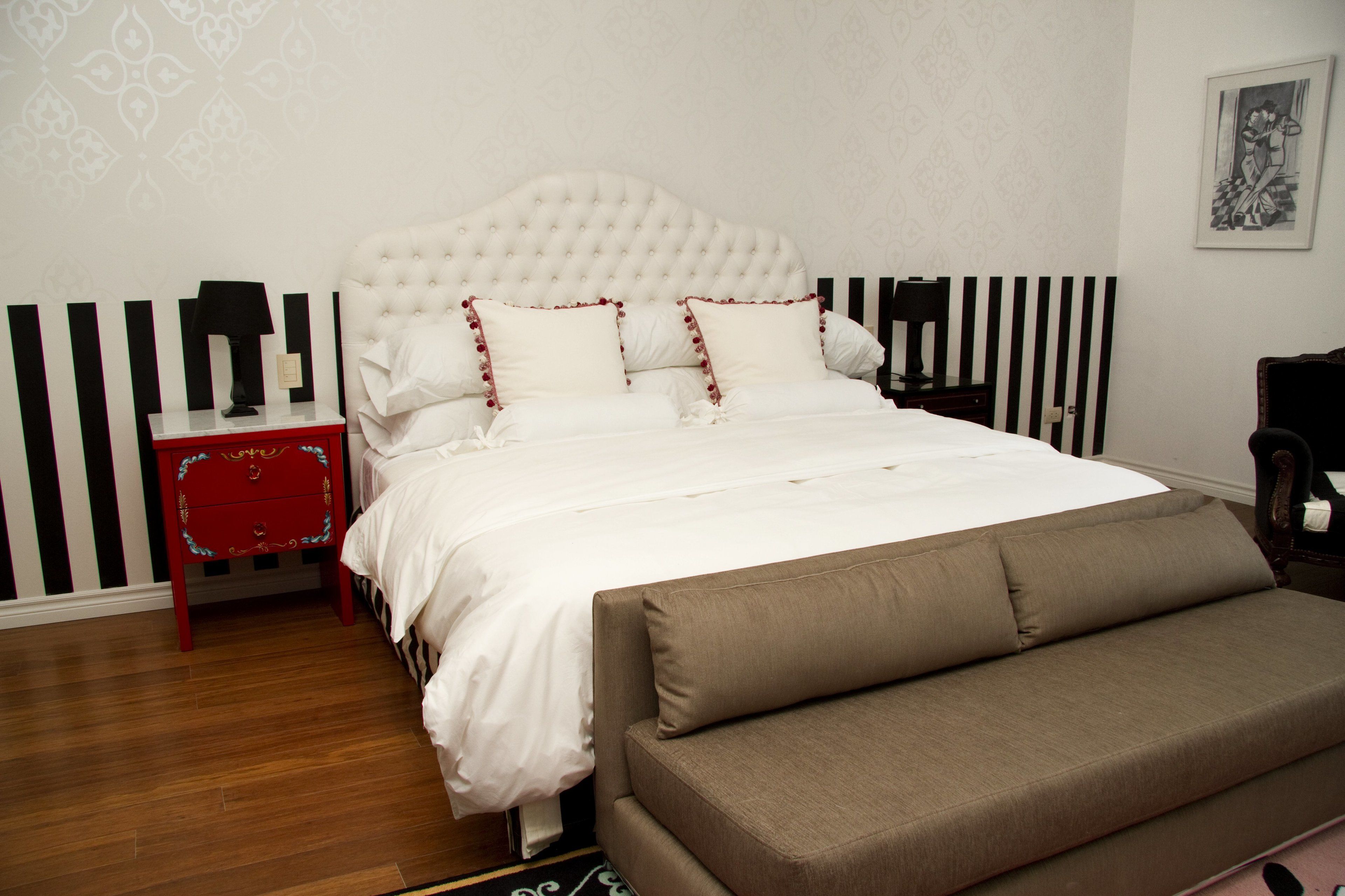 Un dormitorio con cabecera capitonada, cama blanca con almohadas ribeteadas en rojo, mesita de noche roja y sofá marrón.