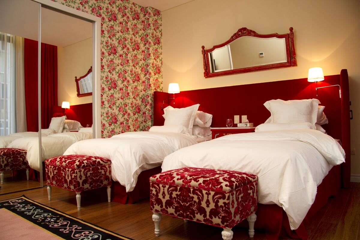 A hotel room with two beds, red and white bedding, matching red and white upholstered chairs, a large mirror, and a floral wallpaper design.