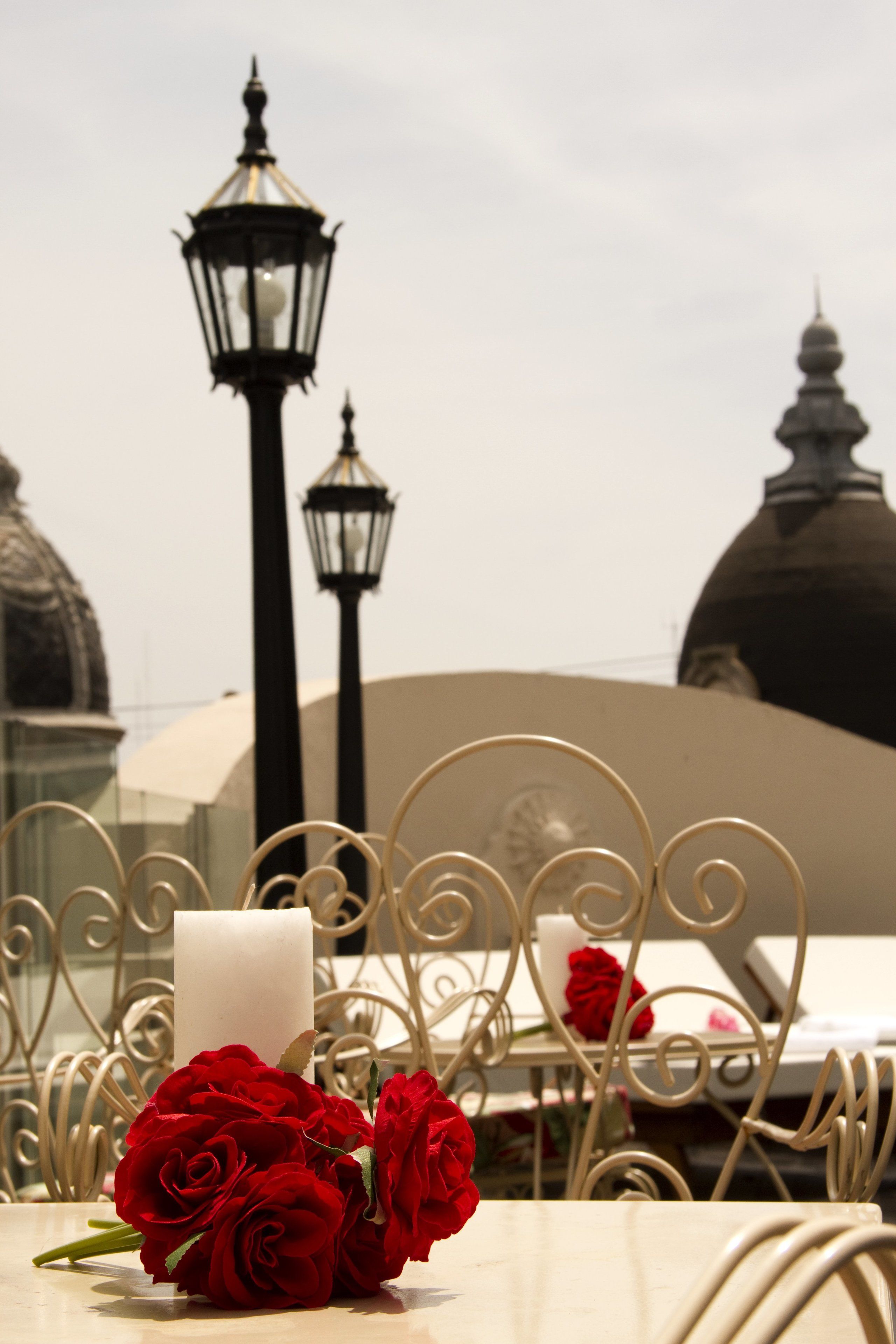 A table with a white candle and red roses, a metal chair, and a view of a building with domes and street lamps.