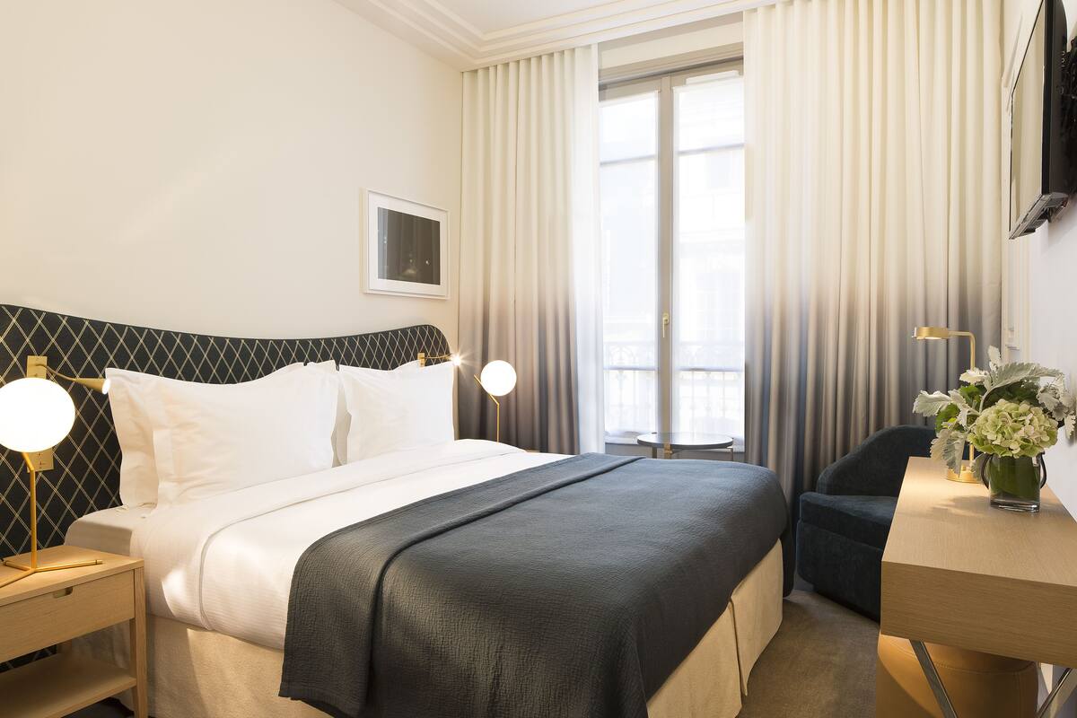 Habitación de hotel con cama, mesitas de noche, escritorio, silla, televisión y ventana con cortinas.