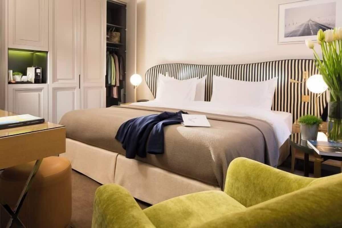 Una habitación de hotel con cama, escritorio, silla, armario y mesita de noche con lámpara.