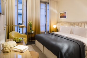Habitación de hotel con cama, una silla, una mesa con libros y una lámpara, una mesita con un frutero, y una ventana con cortinas.