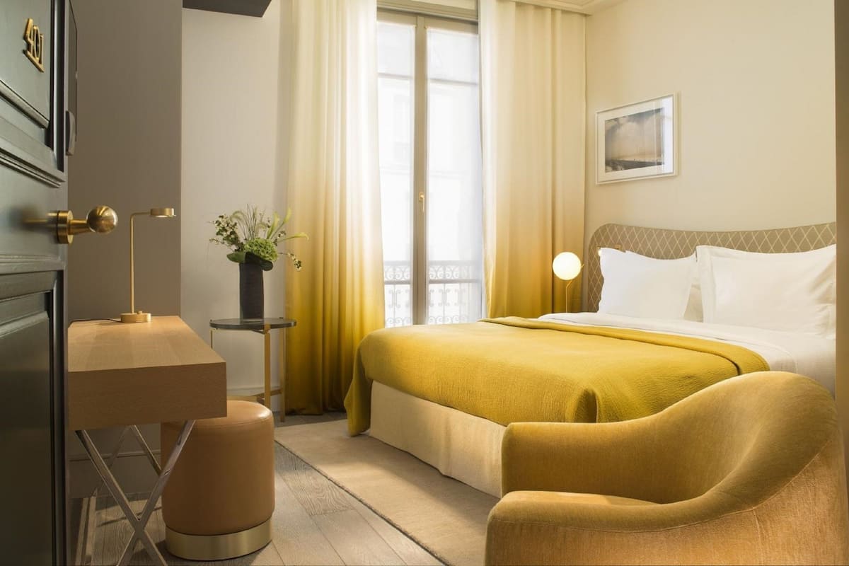 Habitación de hotel con cama, mesita de noche, lámpara, silla, planta en maceta y ventana con cortinas.