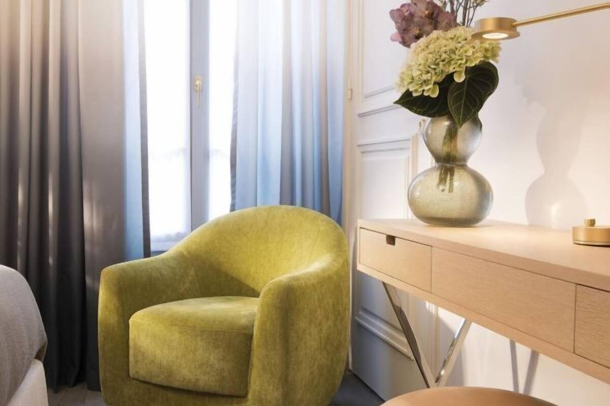 Una habitación con un sillón verde, un escritorio con un florero y una ventana con cortinas.