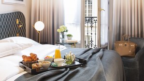 A neatly made bed with a tray of breakfast items, a bedside table with a lamp, and a view of the outside through a window with curtains. - Hôtel Le Marianne (Paris)