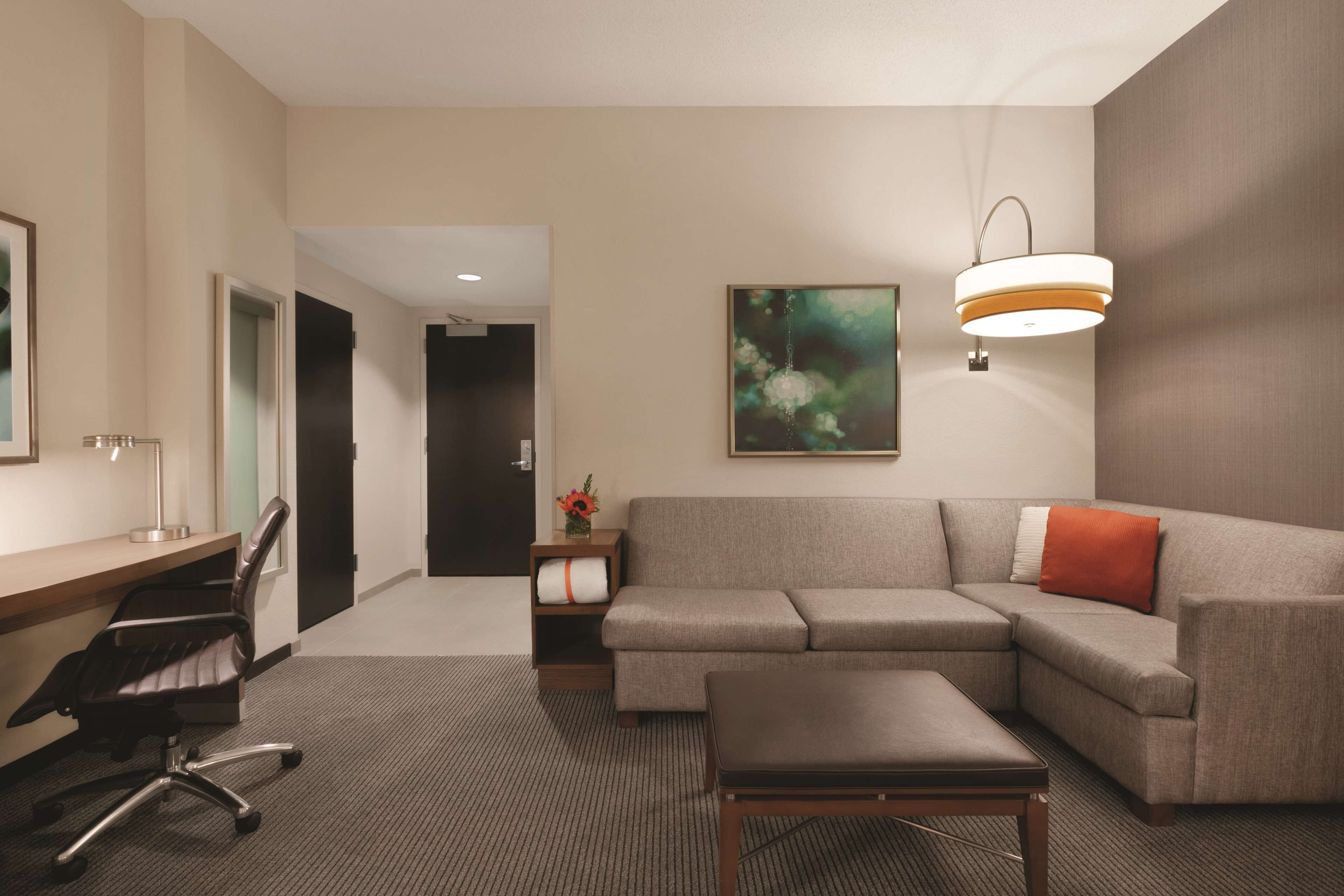 A modern hotel room with a desk, office chair, sofa, and ottoman.
