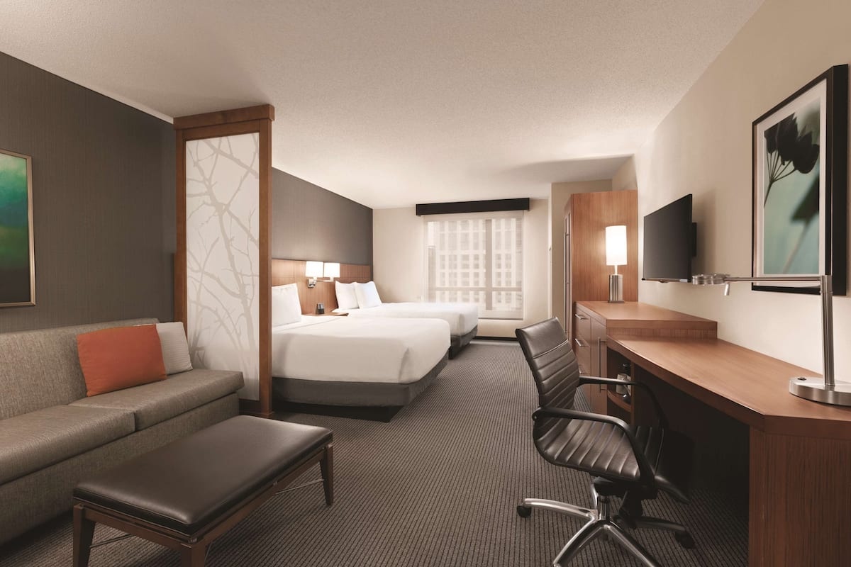 Ein Hotelzimmer mit einer Couch, zwei Betten, einem Schreibtisch, einem Sessel, einem Fernseher und einem Vorhangfenster.