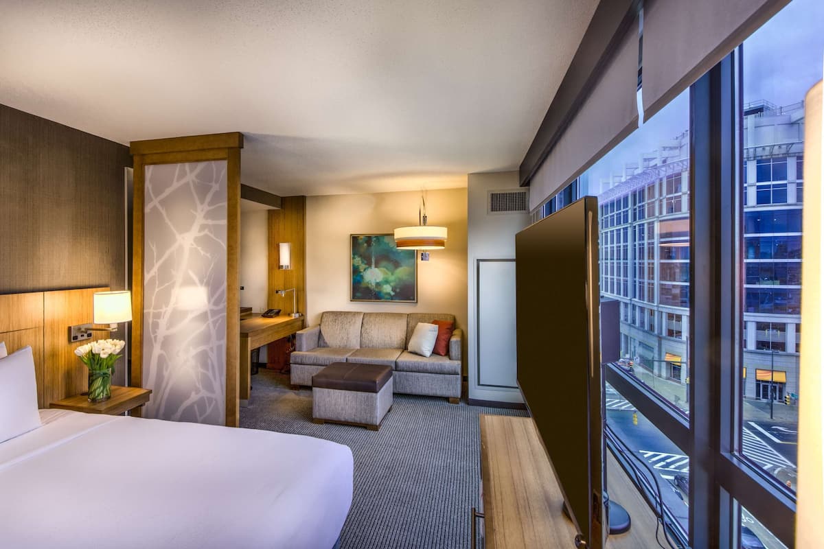 Ein Hotelzimmer mit einem großen Bett, einer Sitzecke, einem Schreibtisch und Blick auf die Stadt durch große Fenster.