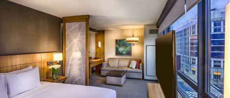 A hotel room with a large bed, a sofa, a desk, and a view of the city through large windows.