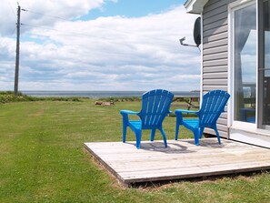 A deck with two blue chairs, a grassy yard, and a view of the ocean. - Cottages on PEI (Chelton)