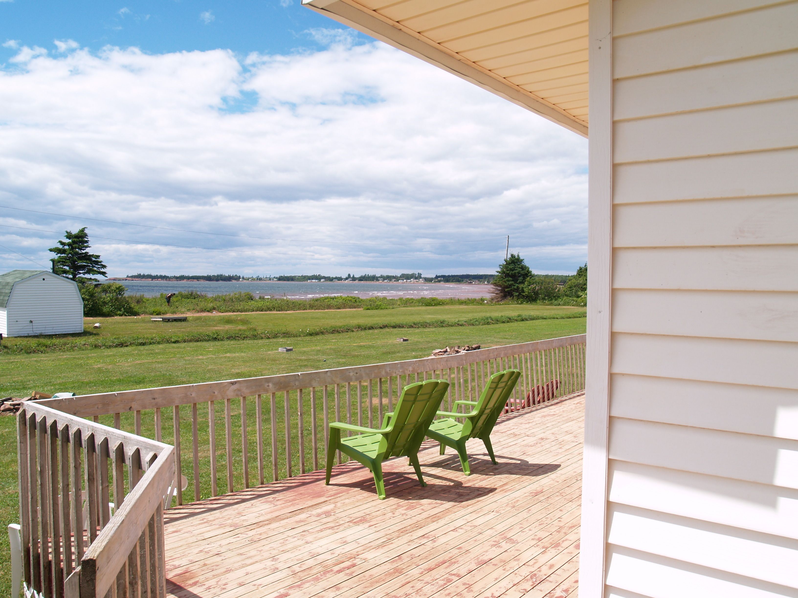 Une terrasse en bois avec deux chaises vertes, un abri de jardin blanc et une vue sur une étendue d’eau et un paysage lointain.