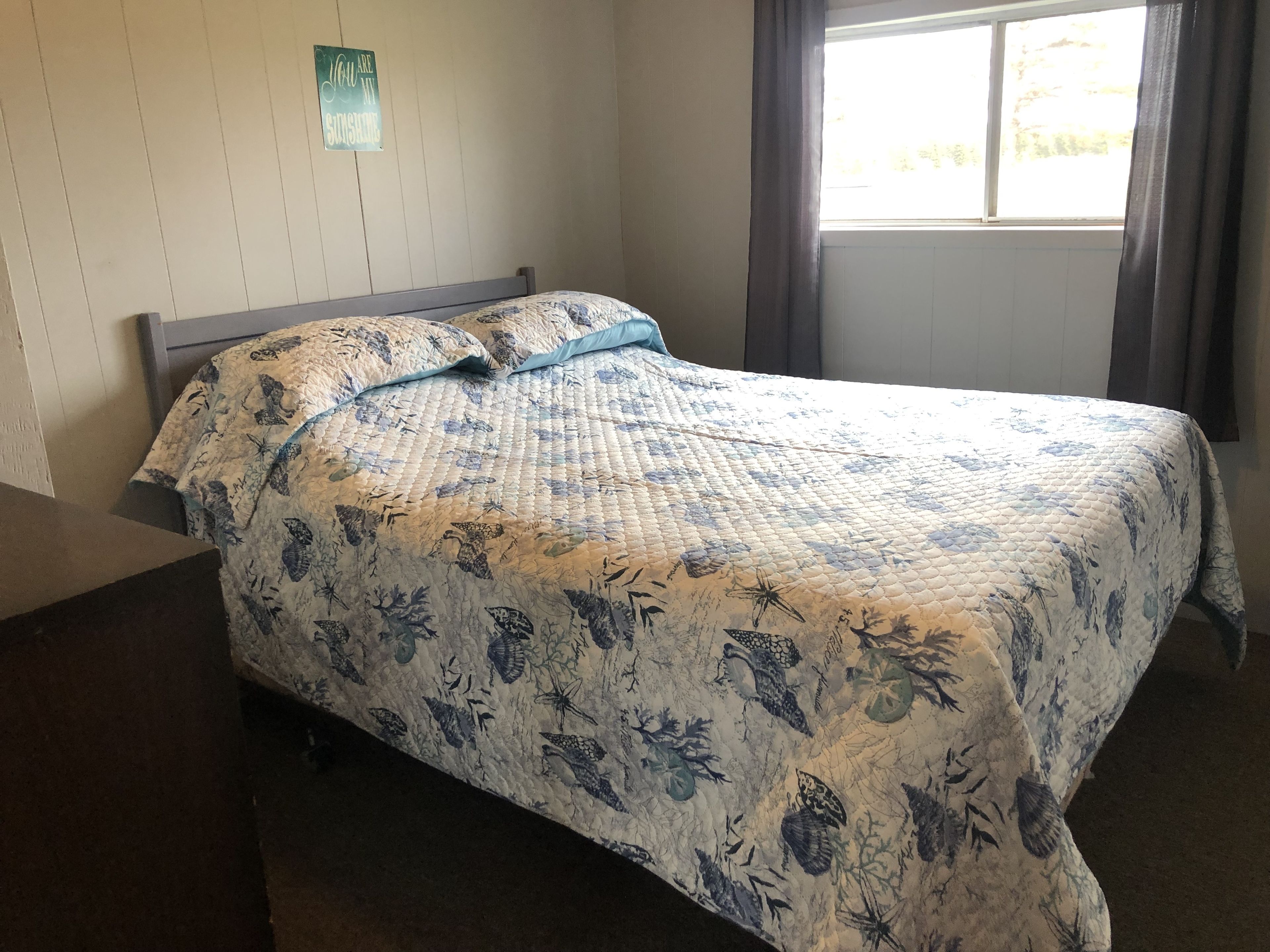 Un lit recouvert d’une courtepointe à motifs floraux, un panneau fixé au mur et une fenêtre avec des rideaux.
