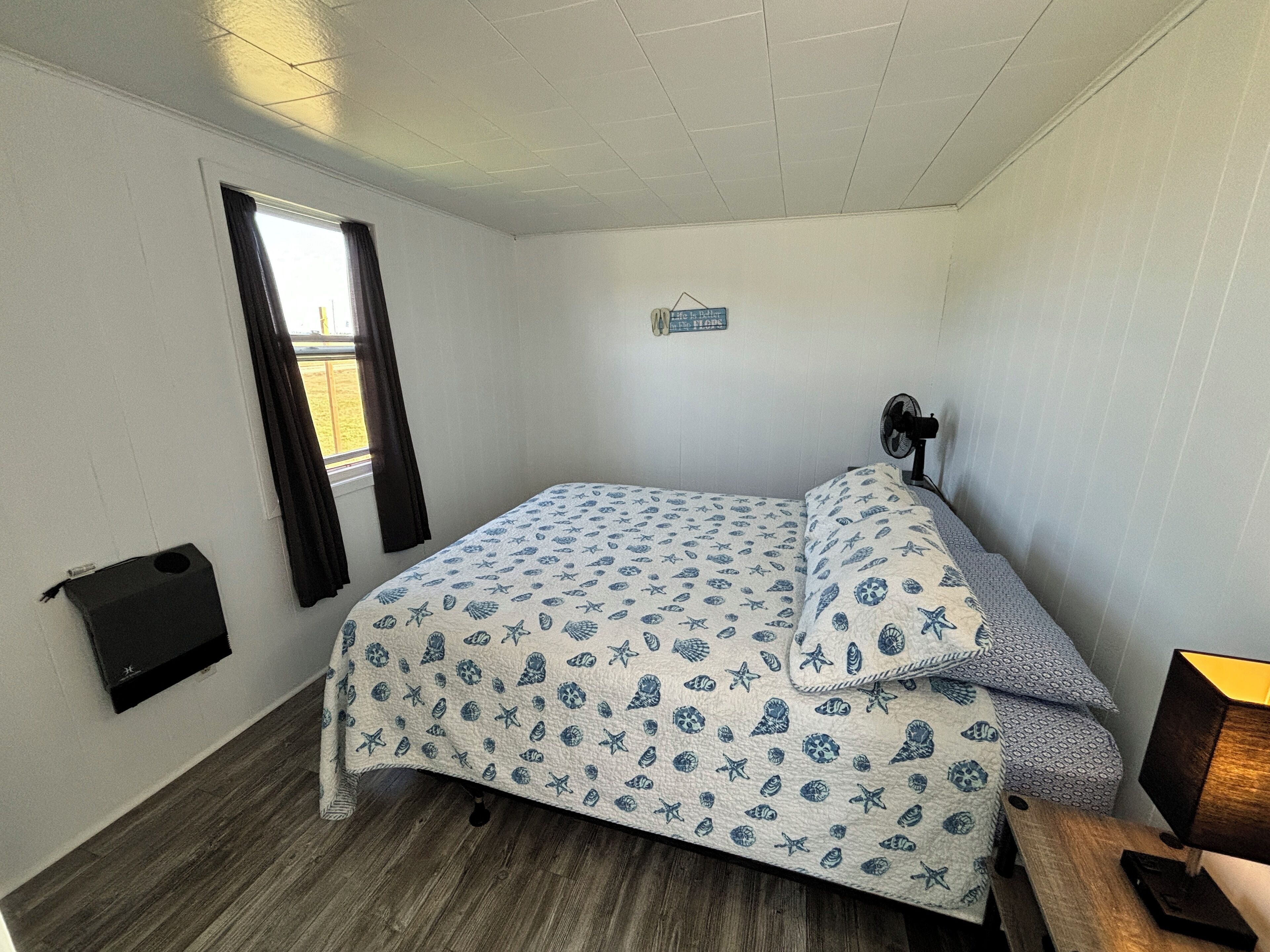 Ein kleines Zimmer mit einem Einzelbett, das mit einem Bezug im maritimen Stil ausgestattet ist, einem Nachttisch mit Lampe, einem Fenster mit Vorhängen und einem an der Wand befestigten Ventilator.