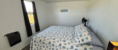 A small, single-bed room with a nautical-themed bedspread, a bedside table with a lamp, a window with curtains, and a wall-mounted fan.