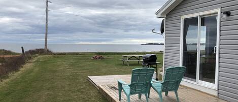 A deck with chairs and a grill, overlooking a grassy area and a body of water.