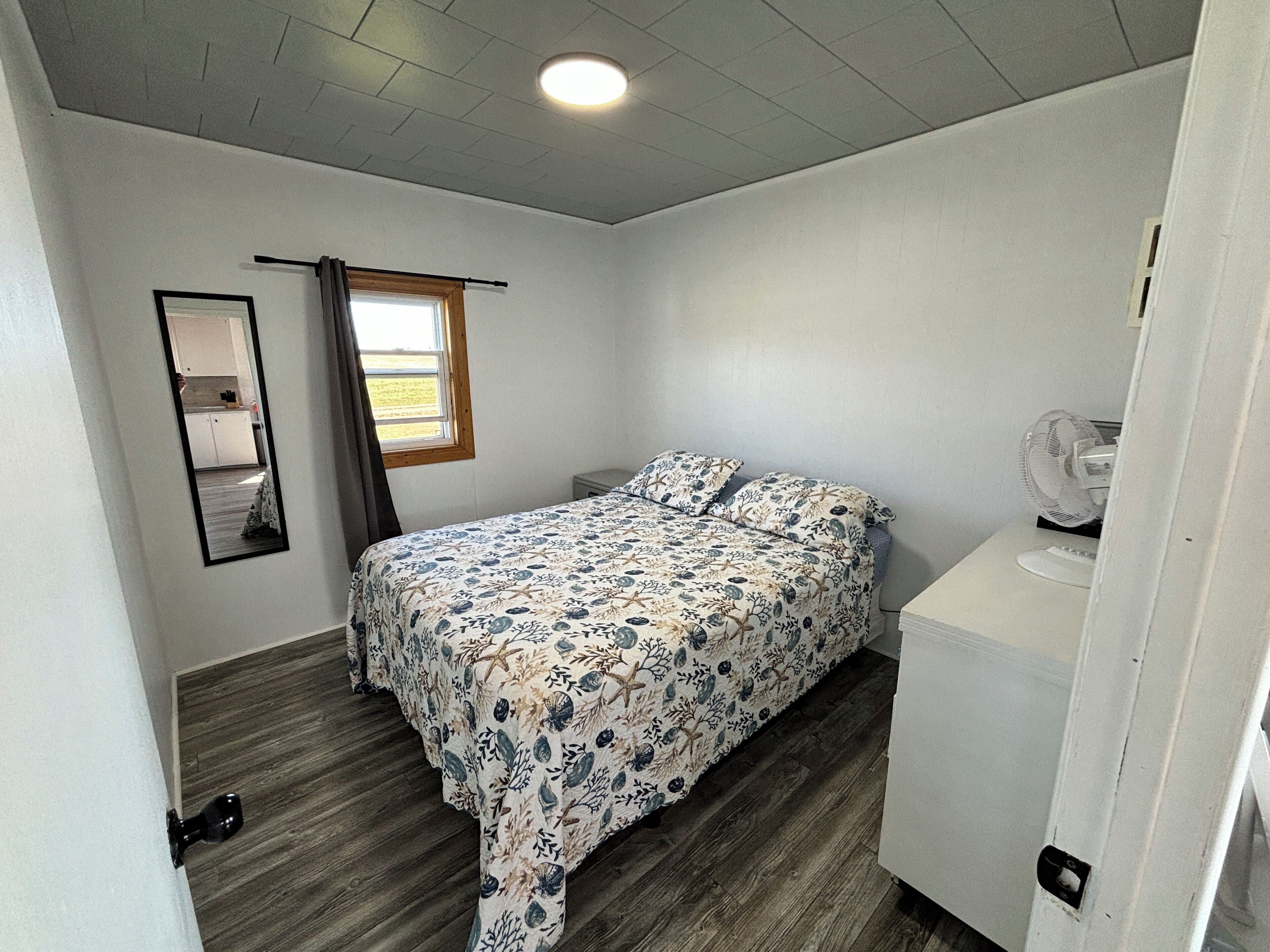 Une chambre à coucher avec un lit, un miroir, une fenêtre avec des rideaux, un ventilateur et une porte.