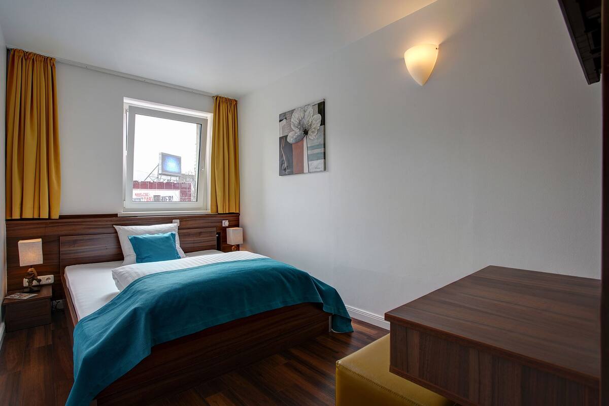 A hotel room with a bed, bedside tables, a window with curtains, a wall-mounted lamp, and a painting.