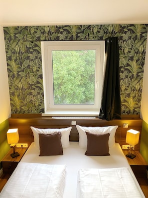 A hotel room with a bed, two bedside tables with lamps, a window with a view of greenery, and a wall with a leafy pattern. - Hotel North (Hamburg)