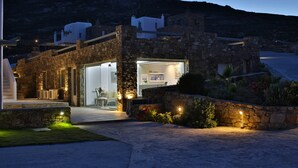 A stone building with a glass entrance, a stone wall with plants, and a view of the surrounding landscape. - Niriides Homes & Villas (Mykonos)