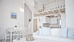 A spacious living area with a white sofa, dining table, and chairs. A staircase leads to an upper level with a balcony. - Niriides Homes & Villas (Mykonos)