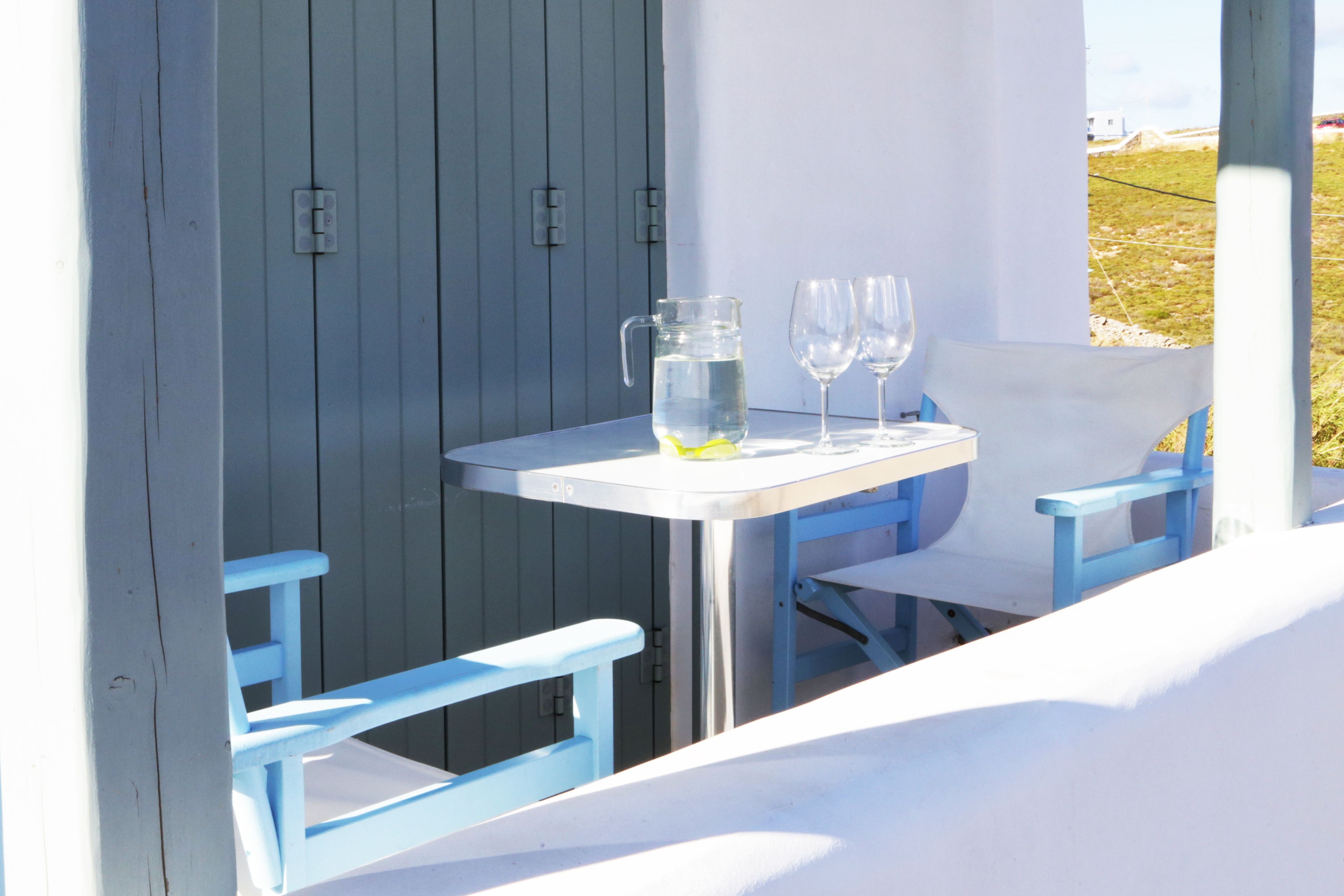 A white table with a pitcher of water and two wine glasses, blue chairs, and a view of a grassy field.