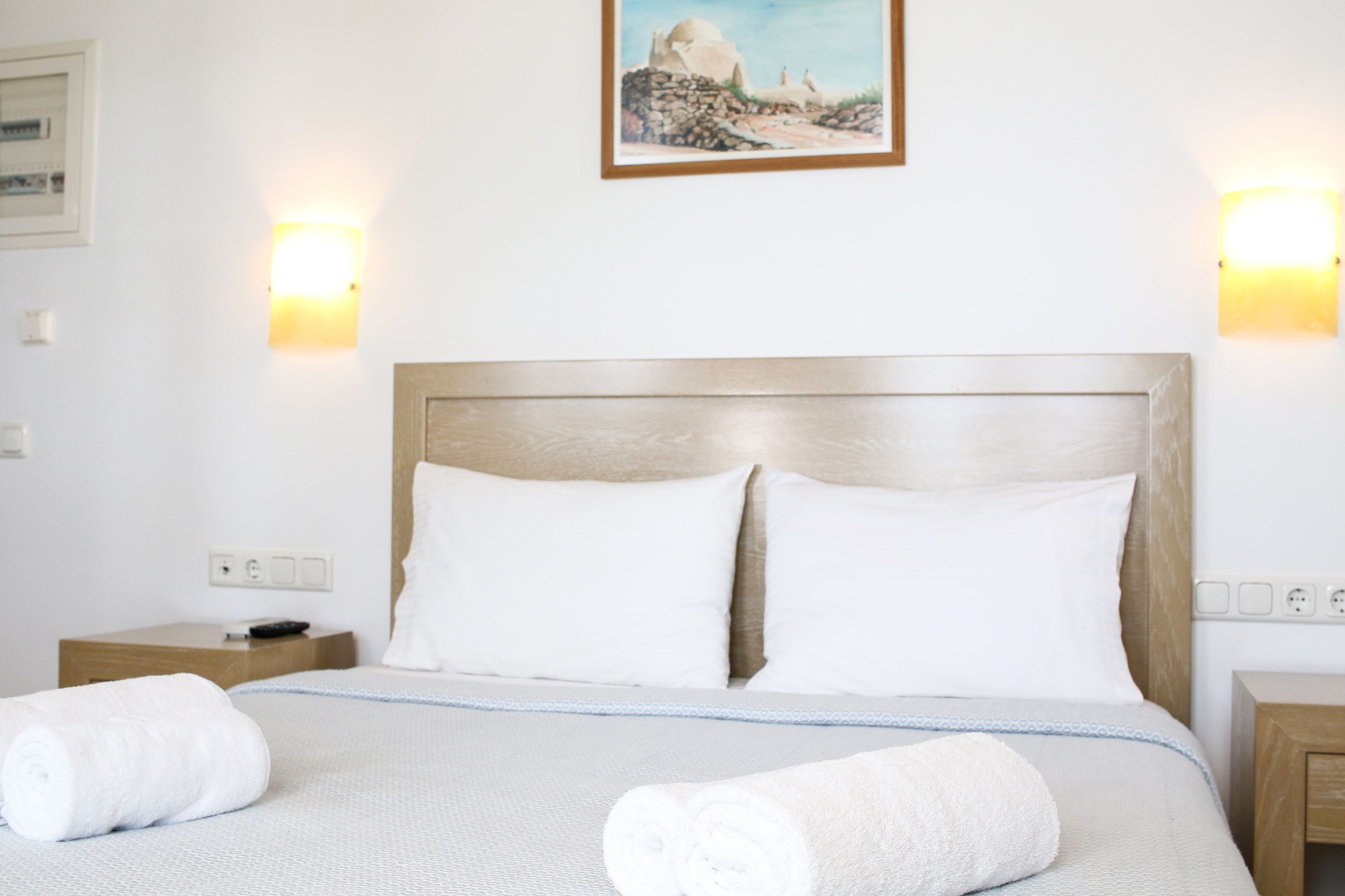 A hotel room with a bed, two bedside tables, a painting on the wall, and wall-mounted lights.