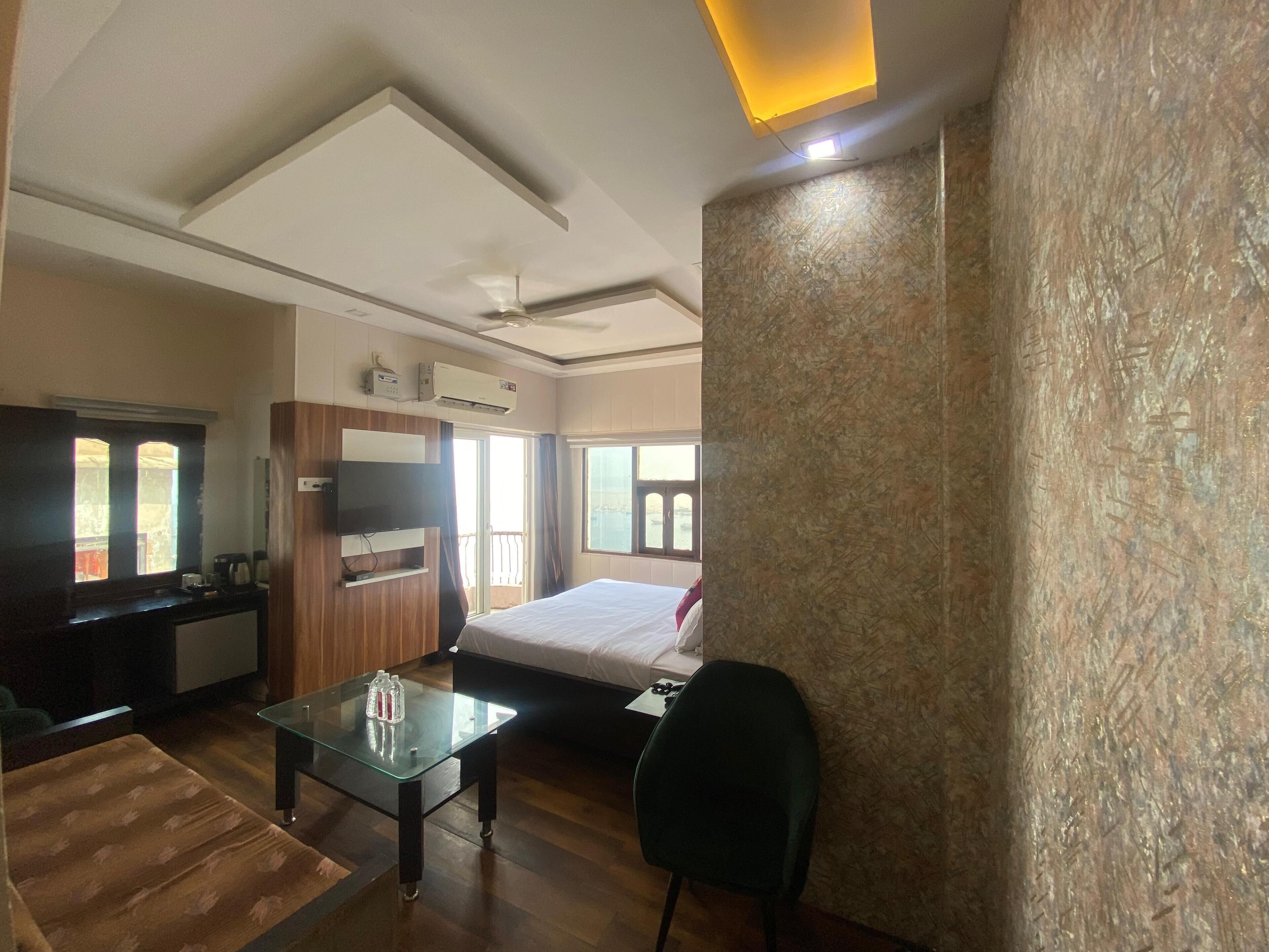 A hotel room with a bed, a glass table with a bottle, a chair, a TV, and an air conditioning unit.