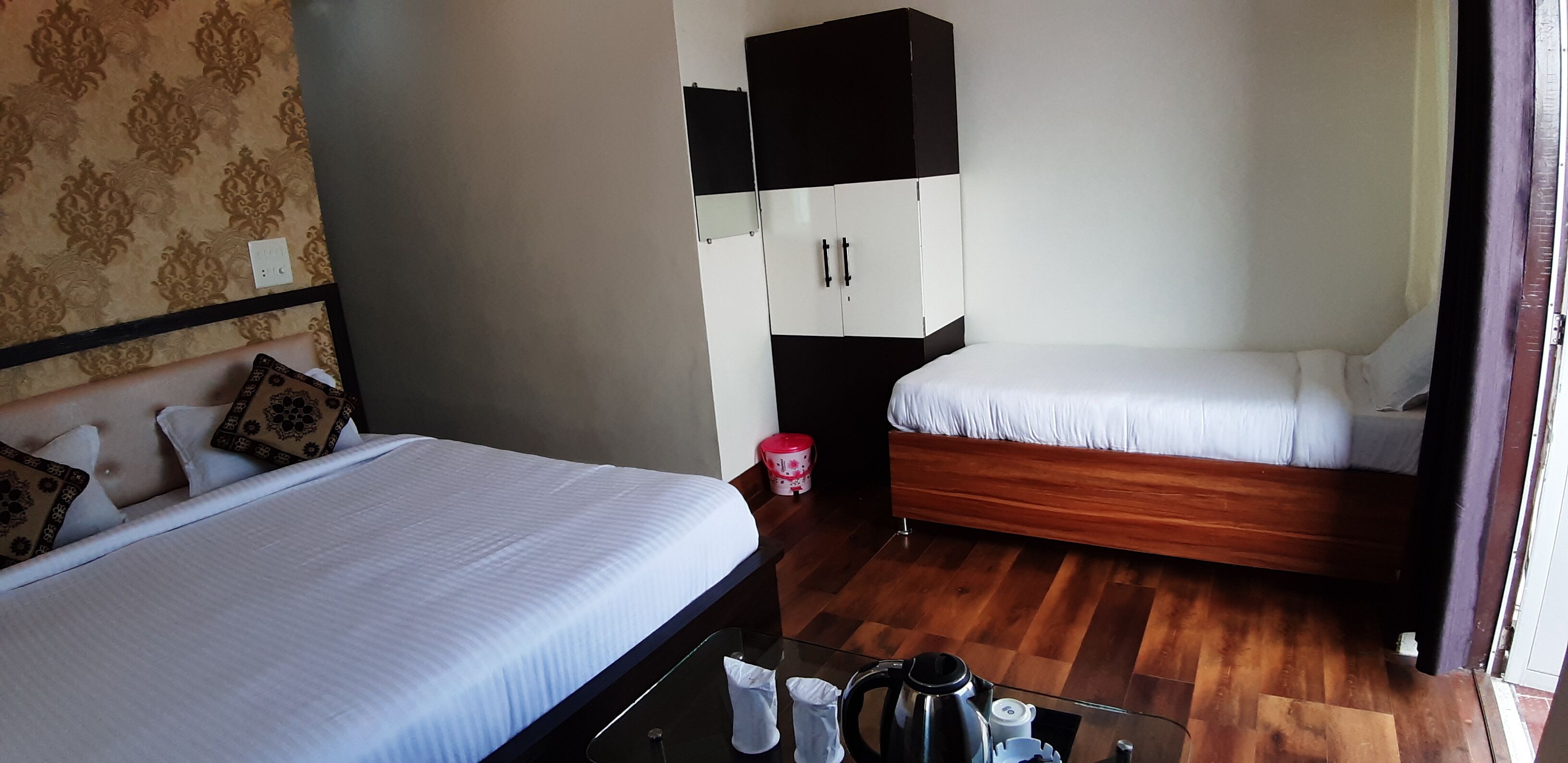 A hotel room with two beds, wooden flooring, a wardrobe, and a small table with a kettle and cup.