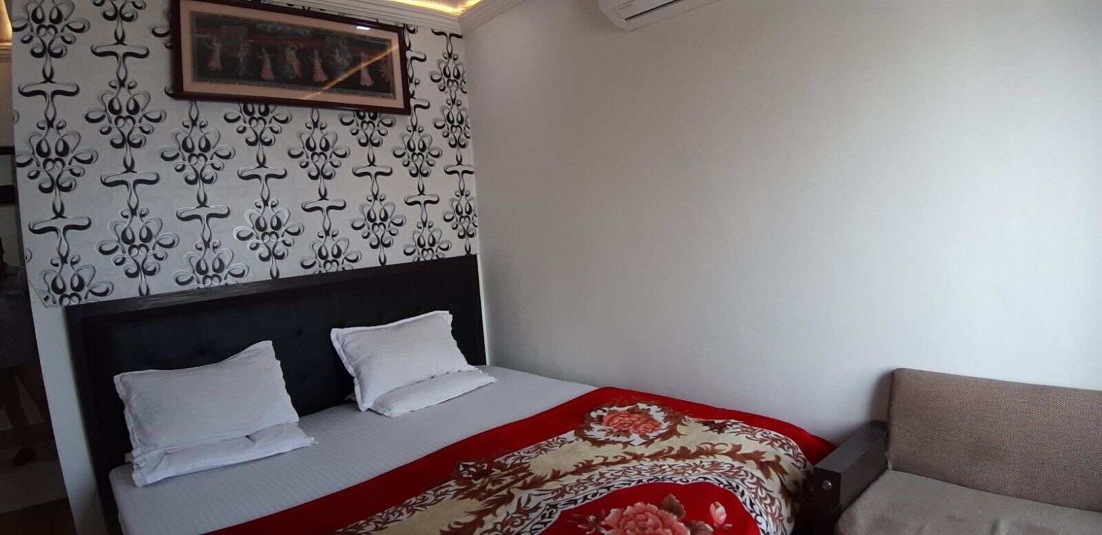 Ett hotellrum med en säng, en sänggavel, ett rött och beige mönstrat sängöverkast, en inramad tavla på väggen och en soffa.