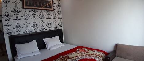 Ett hotellrum med en säng, en sänggavel, ett rött och beige mönstrat sängöverkast, en inramad tavla på väggen och en soffa.