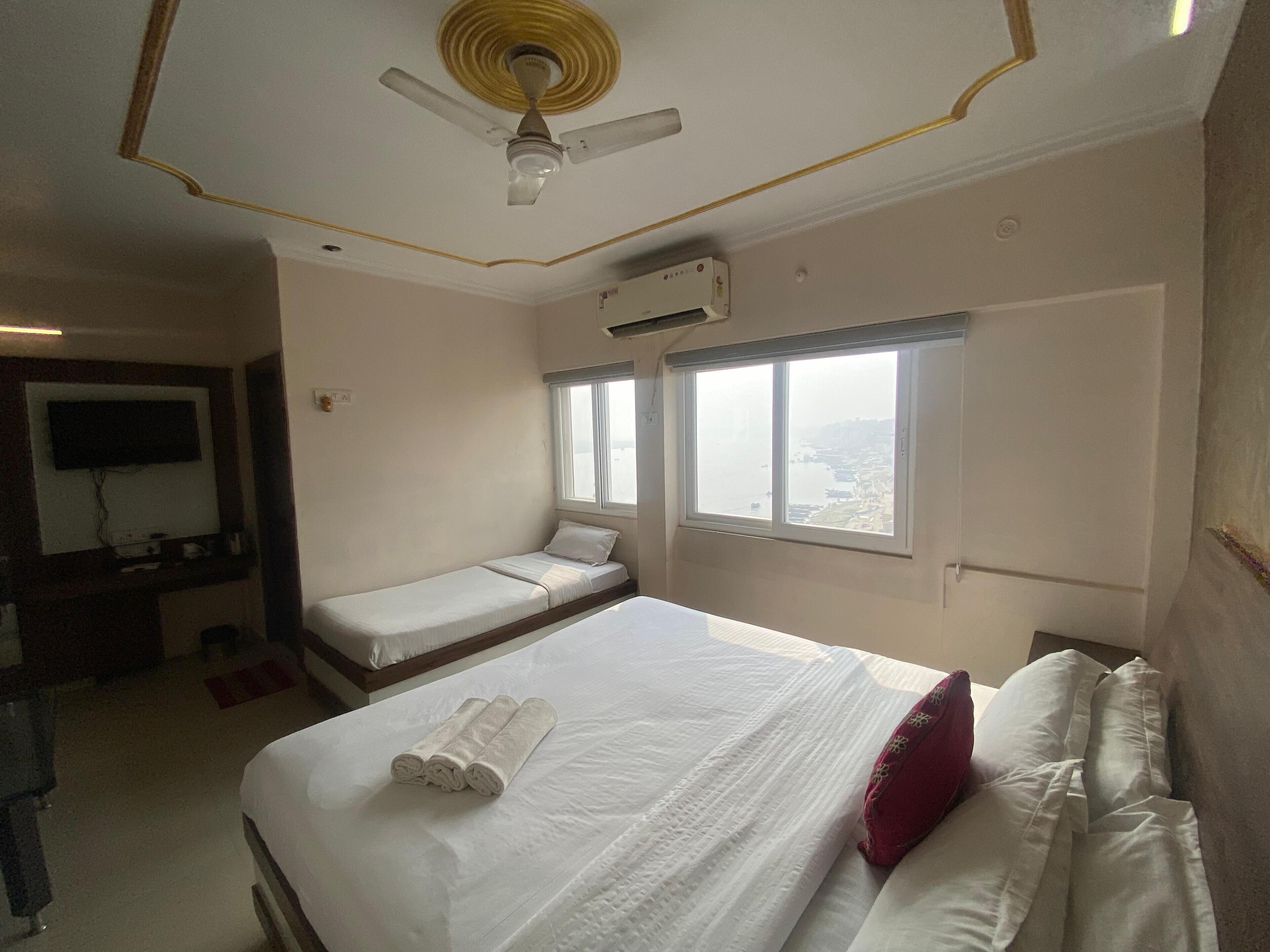 A hotel room with a large bed, a smaller bed, a ceiling fan, an air conditioning unit, a window with a view, and a television on the wall.