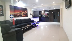 A modern living room with a black leather sofa, a coffee table, and a flat-screen TV. There is a reception desk with a computer and a person sitting behind it. The room has a tiled floor and a ceiling fan. - Hotel Sita (Varanasi)