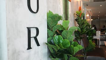 Un área de recepción con una pared de mármol que muestra la palabra "AURA" en letras negras, plantas en macetas y un adorno naranja colgante.