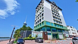 Et moderne hotel med flere etager, med altaner på hver etage, et markant tårn i baggrunden og en parkeringsplads med flere biler.