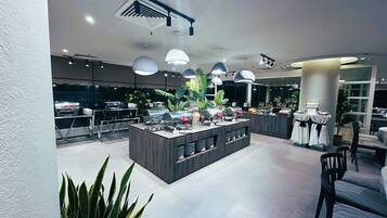 A modern hotel lobby with a central island featuring a buffet setup, surrounded by seating areas and potted plants.
