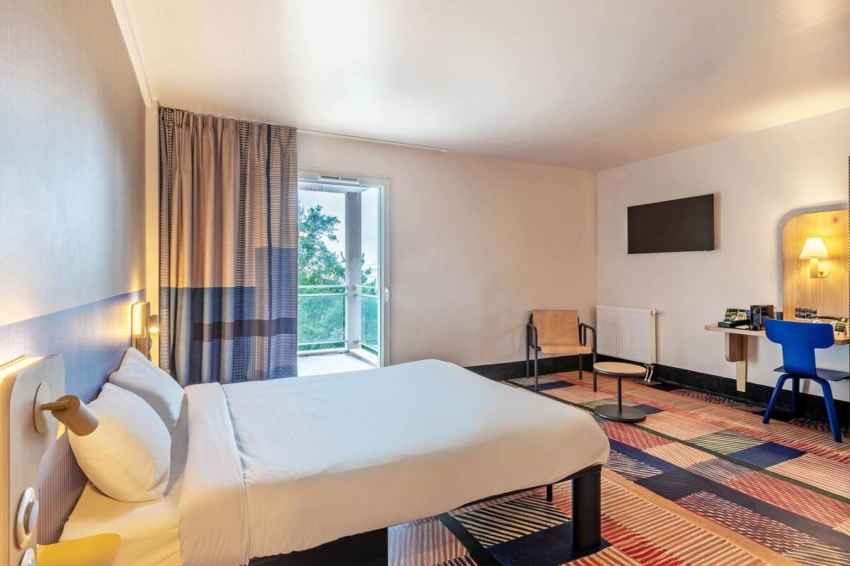 A hotel room with a bed, a desk with a chair, a TV, and a window with curtains.