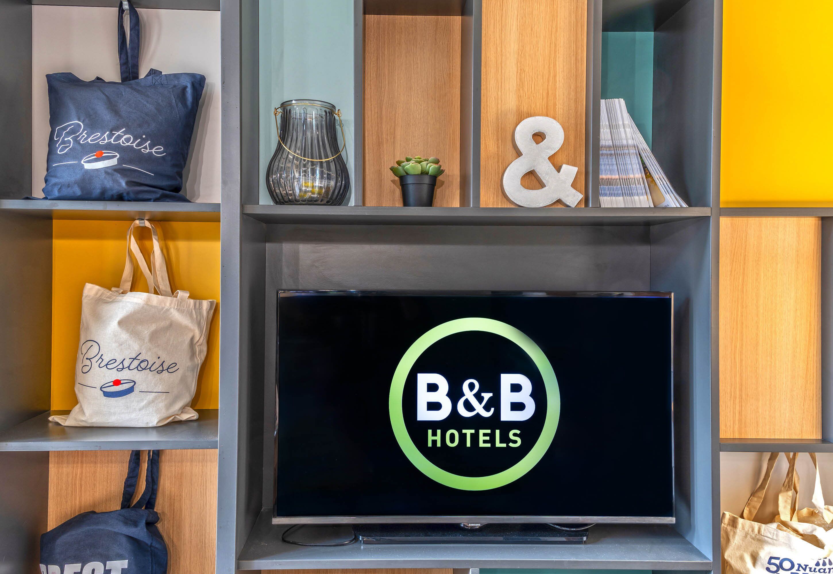 Une étagère avec un panneau indiquant « B&B Hotels », un sac fourre-tout bleu, un sac fourre-tout blanc et une petite plante en pot.