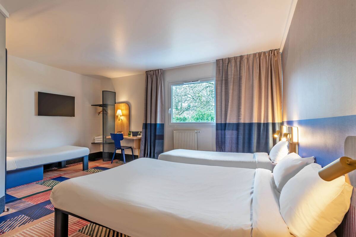 A hotel room with two beds, a desk, a chair, a TV, and a window with curtains.