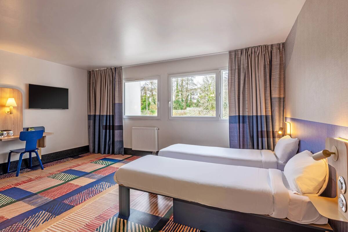A hotel room with two beds, a desk, a chair, and a TV.