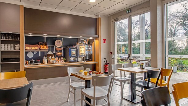 Ein Café mit Tischen und Stühlen, einer Theke mit Speisen und Getränken sowie großen Fenstern mit Blick auf Bäume.
