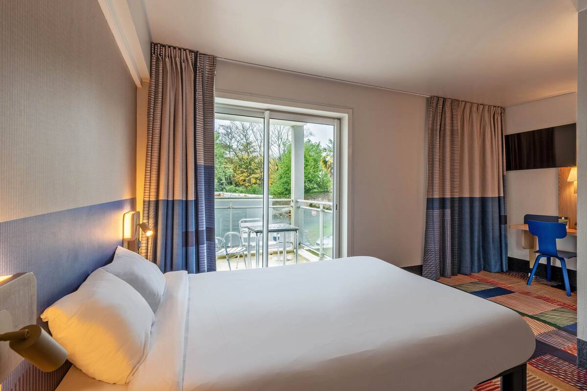 A hotel room with a large bed, a desk with a chair, a balcony with a table and chairs, and curtains.
