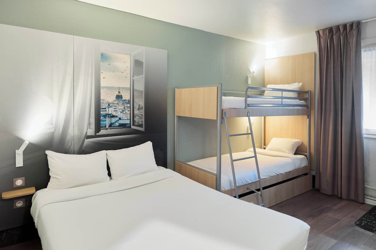 Quarto de hotel com cama grande, beliche, mesa de cabeceira, televisão fixada na parede e janela com cortinas.
