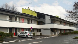 Un hotel de varios pisos con un diseño moderno, que presenta una sección amarilla en el nivel superior, un letrero que dice "Bienvenue" y un área de estacionamiento con un coche blanco.