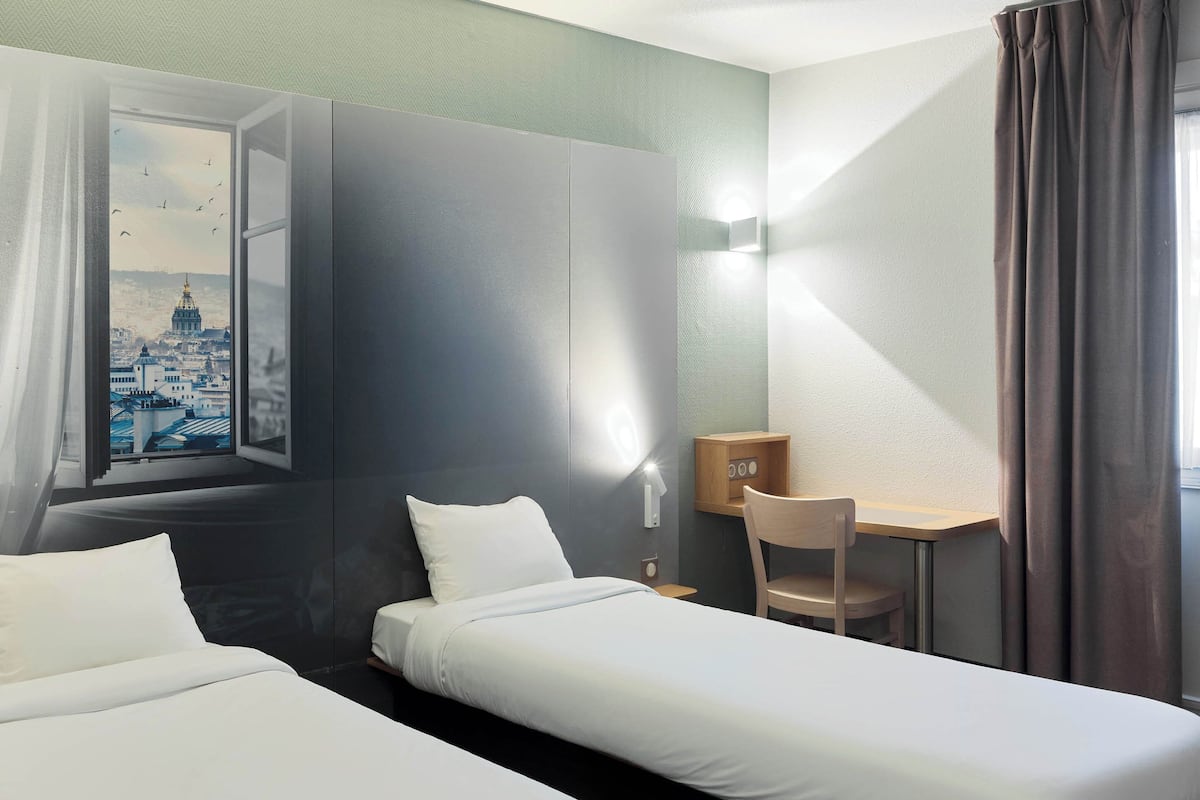 Quarto de hotel com duas camas, uma escrivaninha, uma cadeira, uma janela com vista para a cidade e uma luminária na parede.
