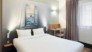 Habitación de hotel con cama, escritorio, silla, ventana con cortinas y una lámpara en la pared.