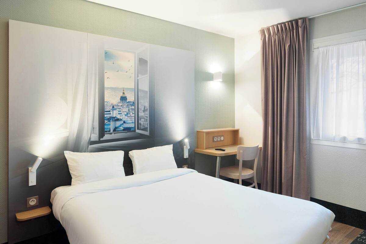 Quarto de hotel com cama, escrivaninha, cadeira, janela com cortinas e luminária na parede.