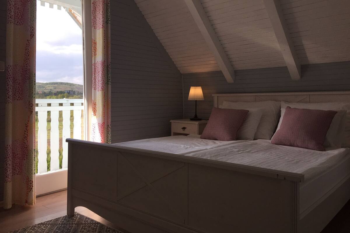 A bedroom with a bed, nightstand, lamp, and a view of the outdoors through a window.