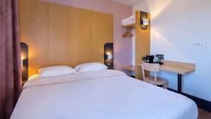 Habitación de hotel con una cama grande, cabecero de madera, lámparas de noche, un pequeño escritorio con una silla y una estantería con toallas.