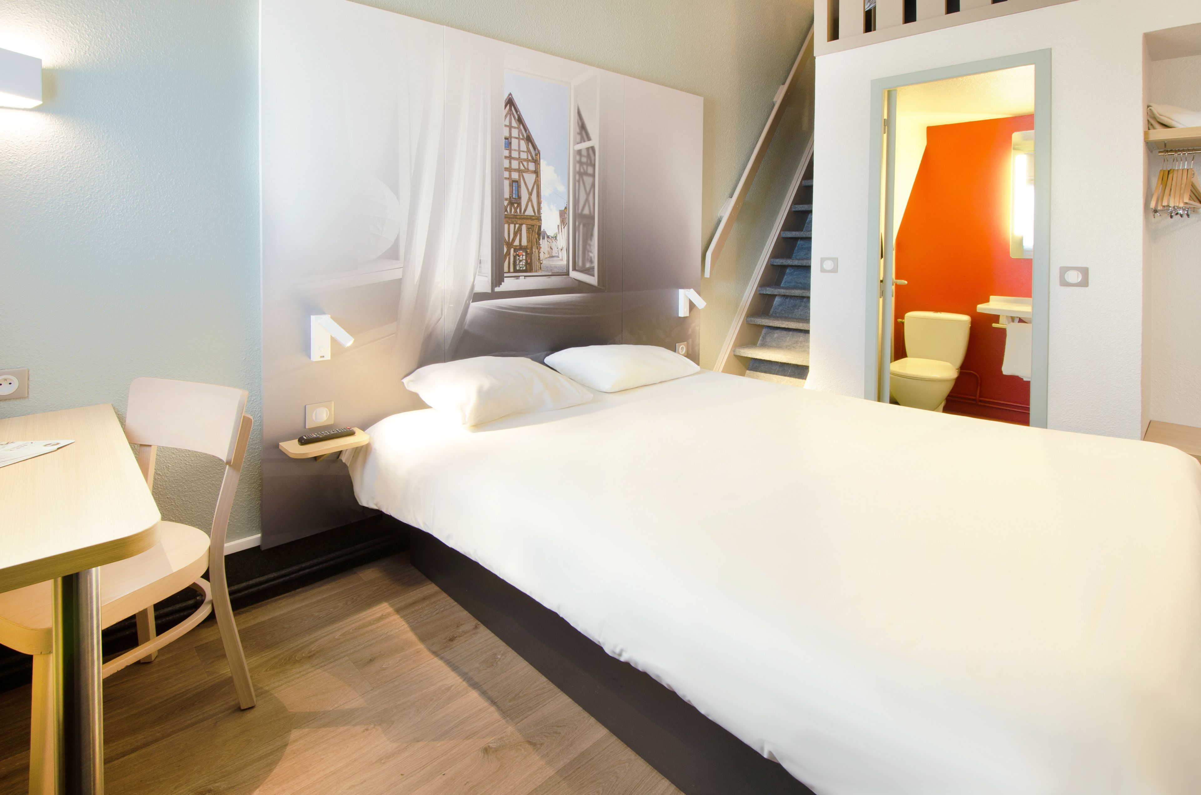 Une chambre d’h ôtel avec un lit, un bureau et une chaise. Un escalier mène à une pièce adjacente.